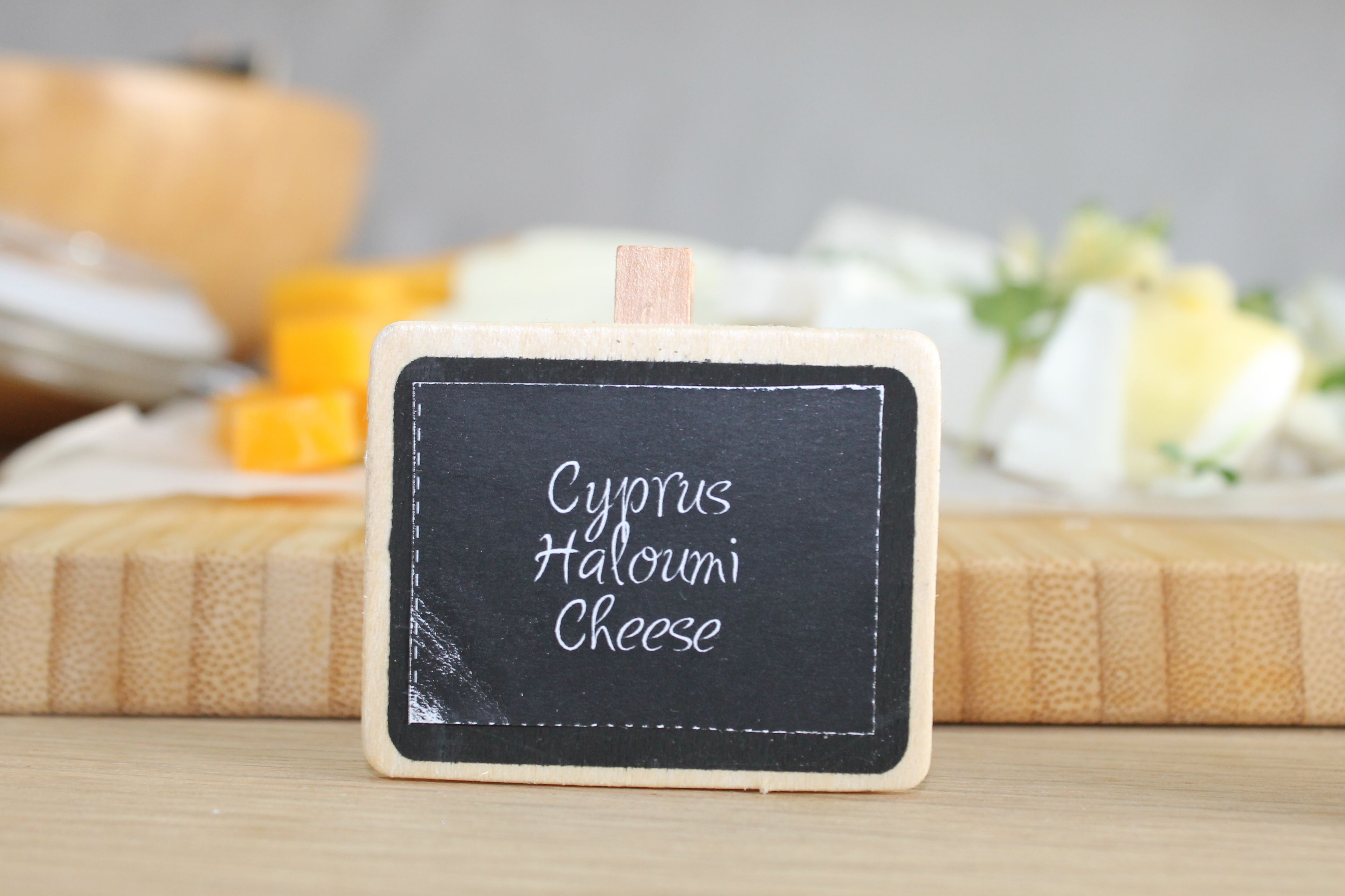 Un pequeño cartel de pizarra con la inscripción 'Cyprus Haloumi Cheese', colocado sobre una superficie de madera, con queso y una calabaza al fondo.