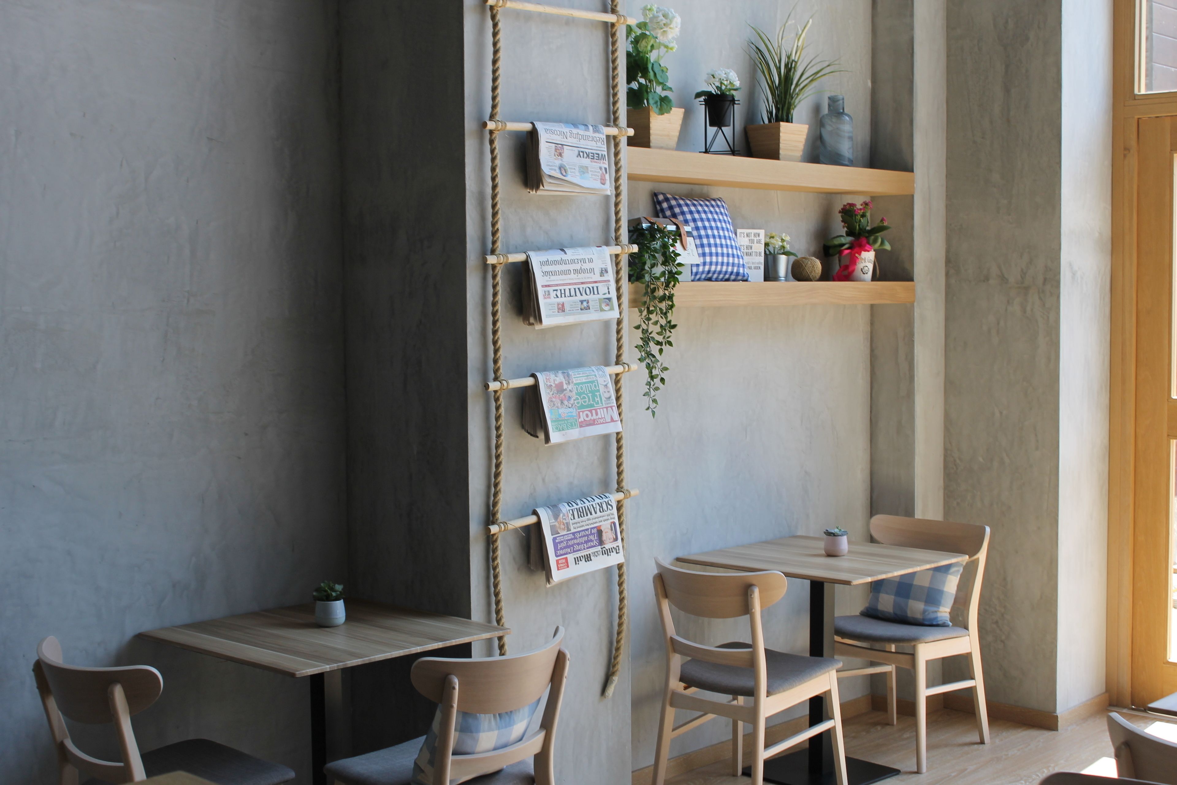 Un café acogedor con mesas y sillas de madera, un estante de cuerda con revistas y plantas en macetas.