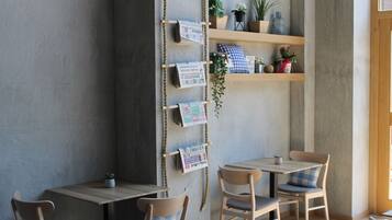 Un café acogedor con mesas y sillas de madera, un estante de cuerda con revistas y plantas en macetas.