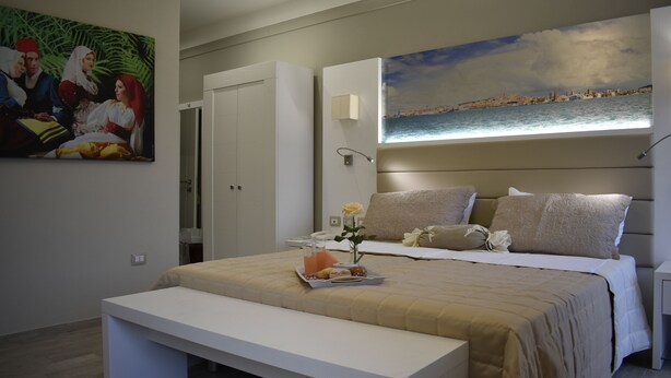 A modern bedroom with a bed, bench, artwork, and a cityscape view. - Il Villino (Cagliari)