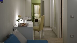 A modern, well-lit room with a blue sofa, a small dining table with a vase of flowers, and a chair. There is a hallway leading to another room with a bed. - Il Villino (Cagliari)