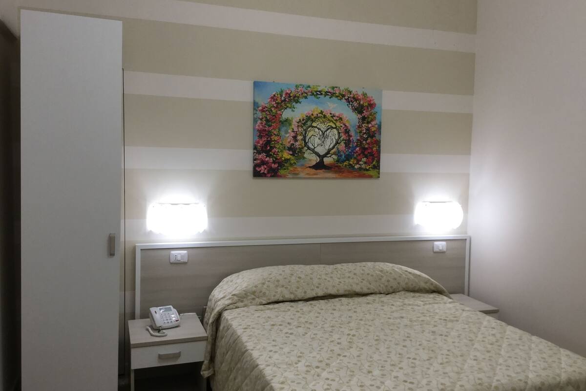 A hotel room with a bed, a nightstand with a phone, a painting on the wall, and two wall-mounted lights.