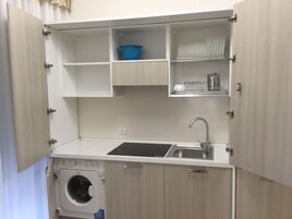 Una kitchenette compacta con lavadora integrada, un lavamanos y estantes para almacenamiento.
