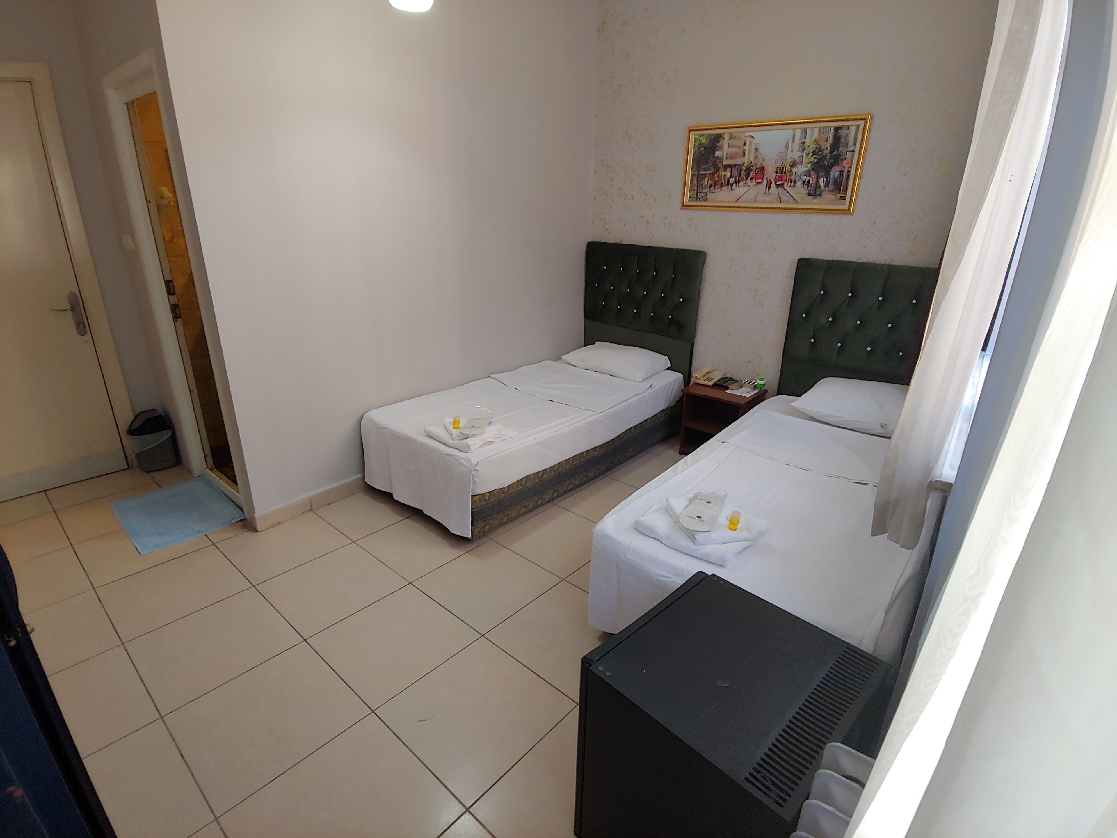 A hotel room with two single beds, a small desk, a trash bin, and a painting on the wall.