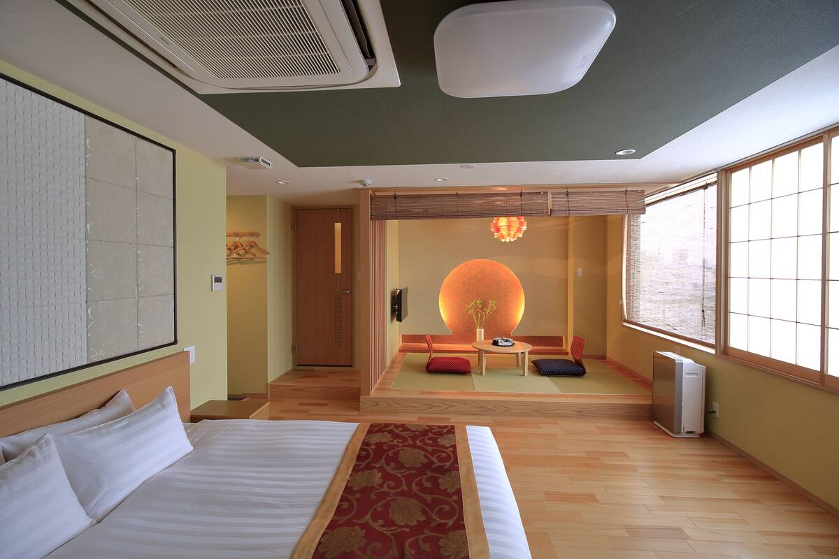 A hotel room with a large bed, a wooden floor, a wall-mounted air conditioner, a ceiling light, and a large window with blinds.