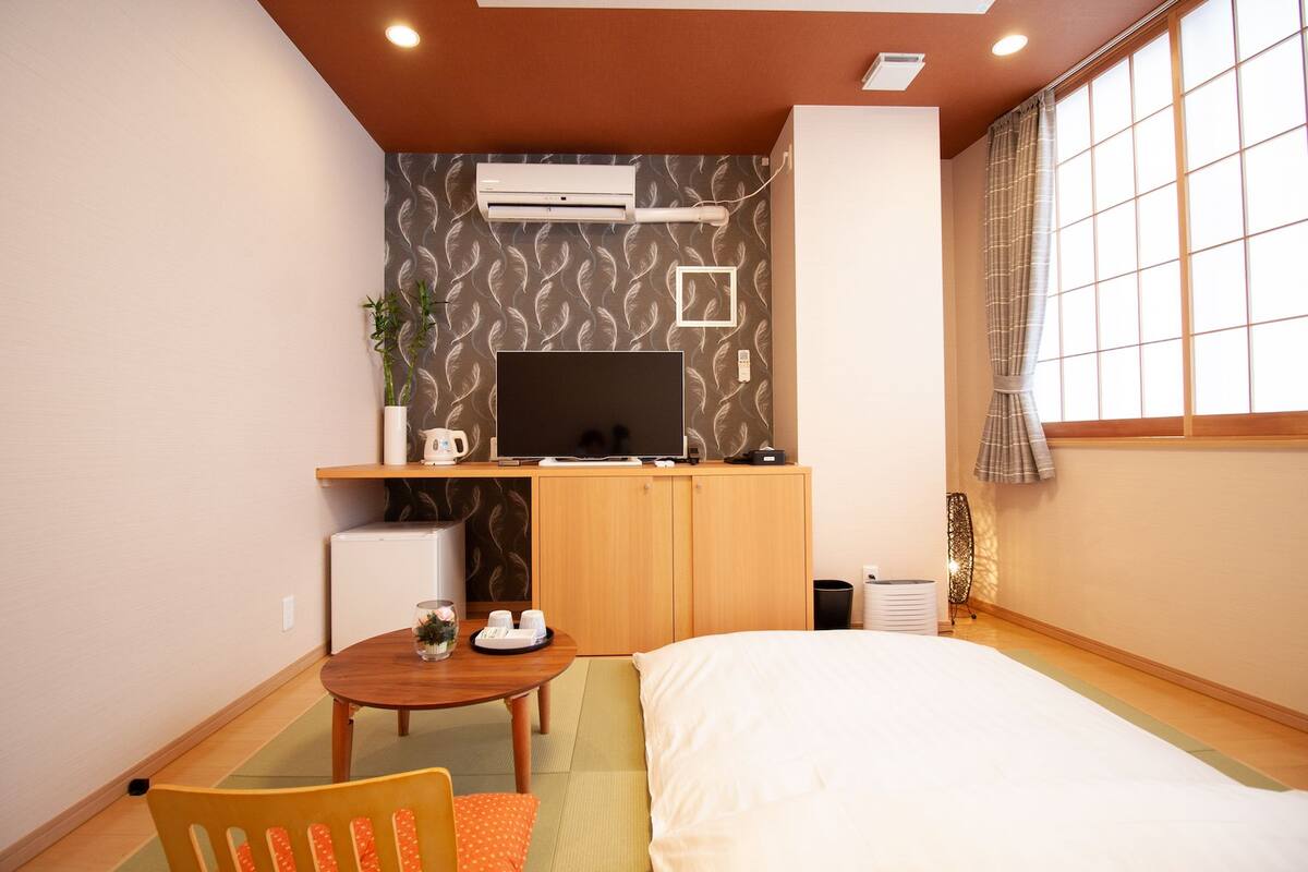 A compact hotel room with a bed, a desk with a TV, a small fridge, a chair, and a window with curtains.