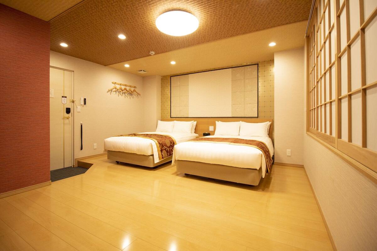 A hotel room with two beds, a wooden floor, a large window with bars, and a ceiling with recessed lighting.