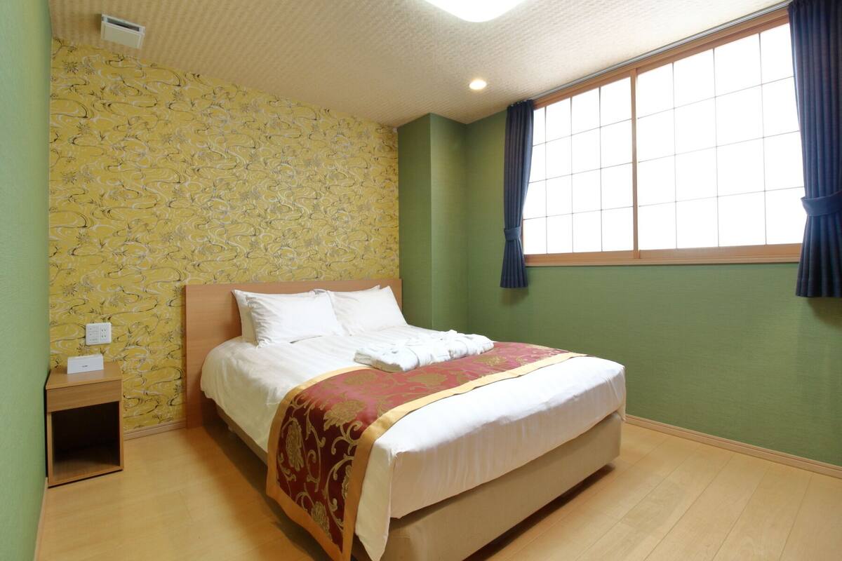 A neatly made bed in a room with a patterned wallpaper, a wooden nightstand, and a window with curtains.
