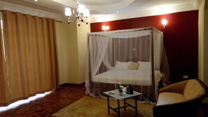 Un dormitorio con una cama con dosel, una mesita con una planta y un hervidor, una silla y un ventanal con cortinas.