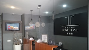 A hotel reception desk with a modern design, a digital screen displaying food, clocks, and a sign for Hotel Kapital. - Hotel Kapital (Maputo)
