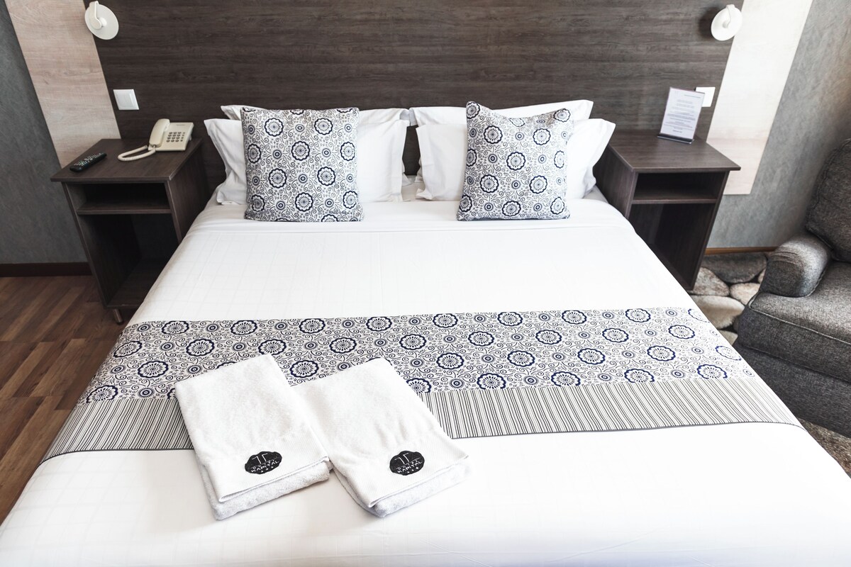 A neatly made bed with patterned pillows and a folded towel, a bedside phone, and a nightstand with a lamp.