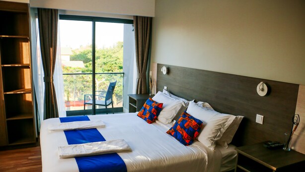 A hotel room with a large bed, a nightstand, a chair, and a view of greenery outside. - Hotel Kapital (Maputo)