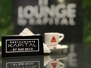 A black sign with white text for 'RESTAURANTE KAPITAL' and a phone number. A white cup with a red triangle logo is on a saucer. - Hotel Kapital (Maputo)