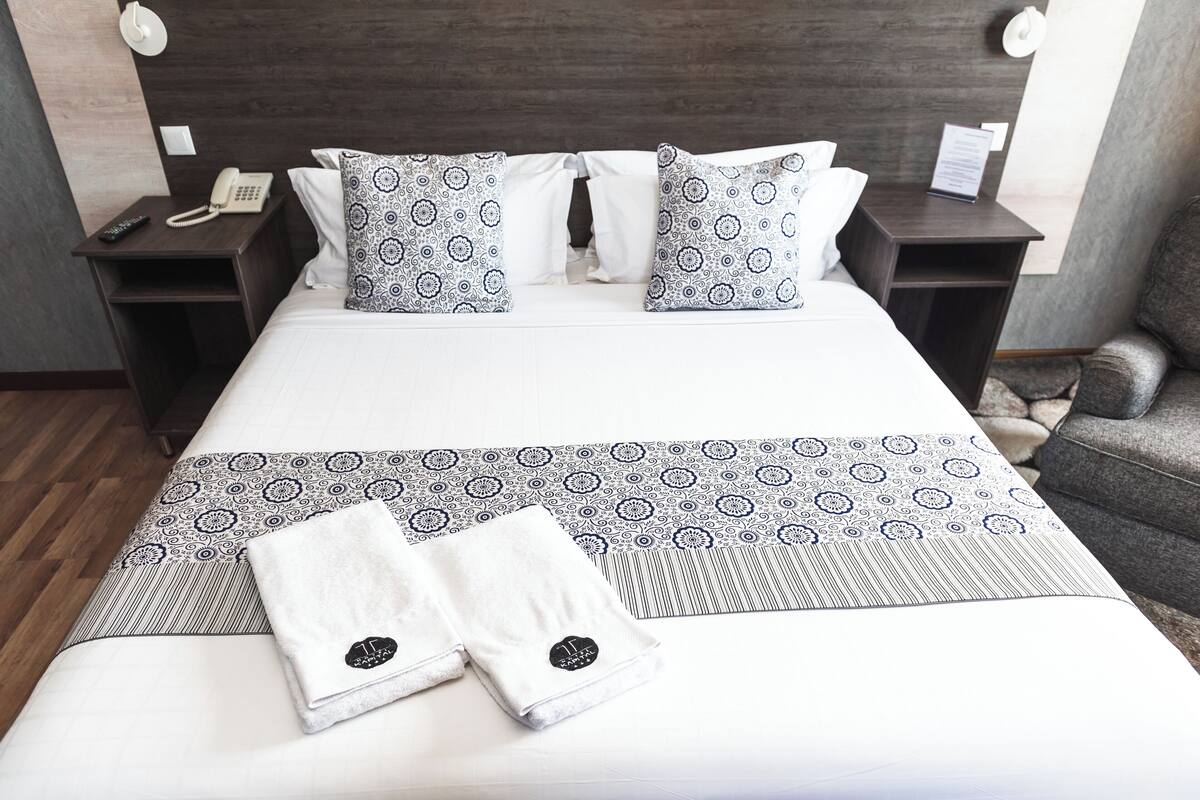 A neatly made bed with patterned pillows and a folded towel, a bedside phone, and a nightstand with a lamp.