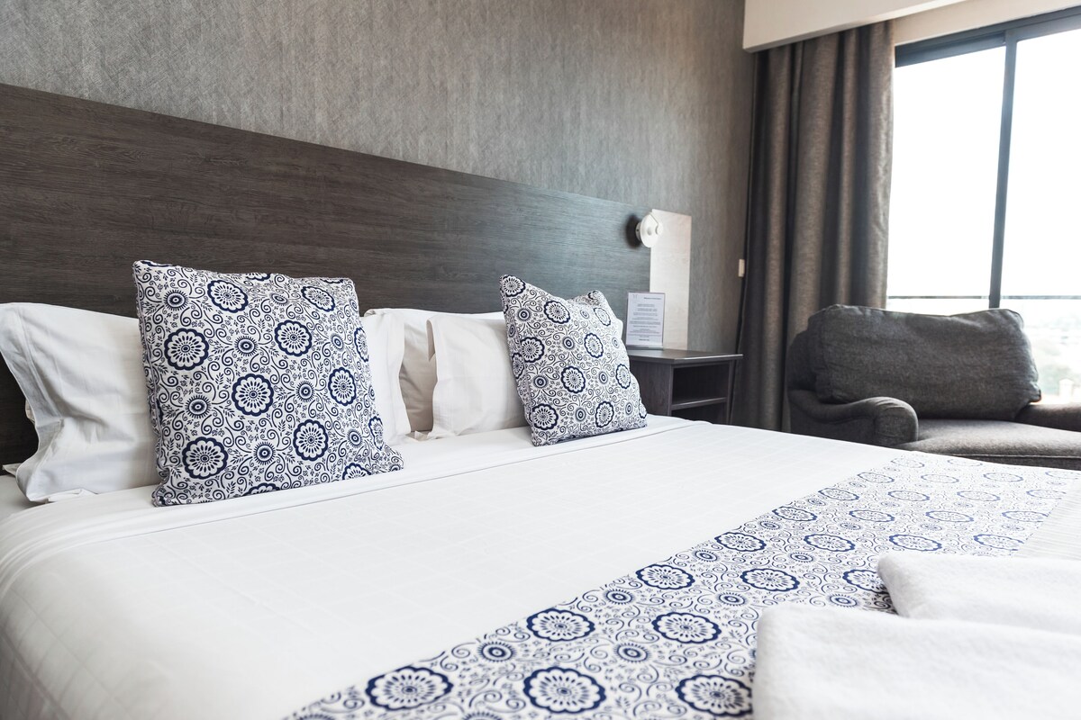 A hotel room with a large bed, two patterned pillows, a grey armchair, and a window with curtains.