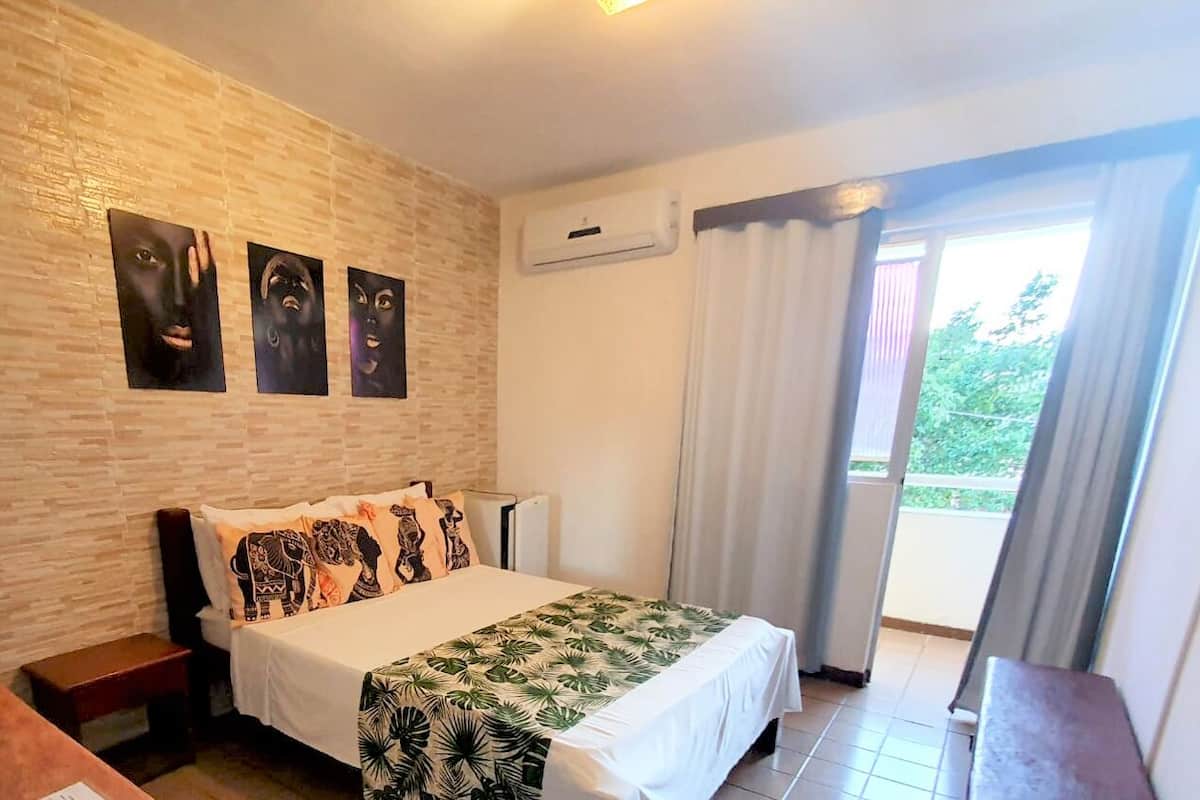 Hotellihuone, jossa on sänky, yöpöytä, puinen penkki, kuvioitu seinä ja ikkuna, jossa on verhot.