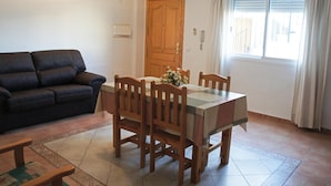 Een woonkamer met een donkerblauwe bank, een houten eettafel met vier stoelen en een bloemenstuk. Er is een houten deur en een airconditioningunit op de muur.