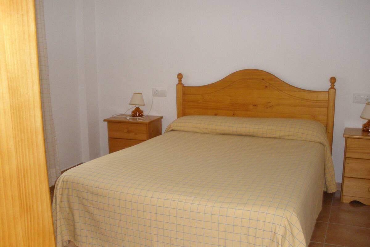 Een enkel bed met houten hoofdeinde in een kamer met witte wanden en betegelde vloer.