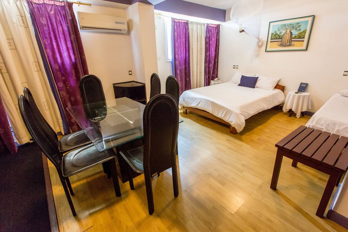 A hotel room with a wooden floor, a glass dining table with chairs, a bed with white bedding, a small table with a white tablecloth, and purple curtains.