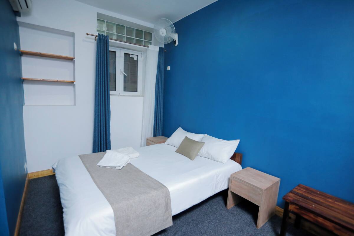 A neatly made bed with white and beige bedding, a bedside table, a wooden bench, and a window with blue curtains.