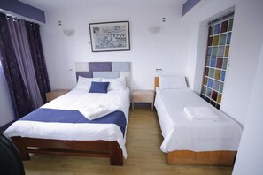 A hotel room with two single beds, a wooden headboard, a small bedside table, a nightstand, a window with a colorful view, and a framed picture on the wall.