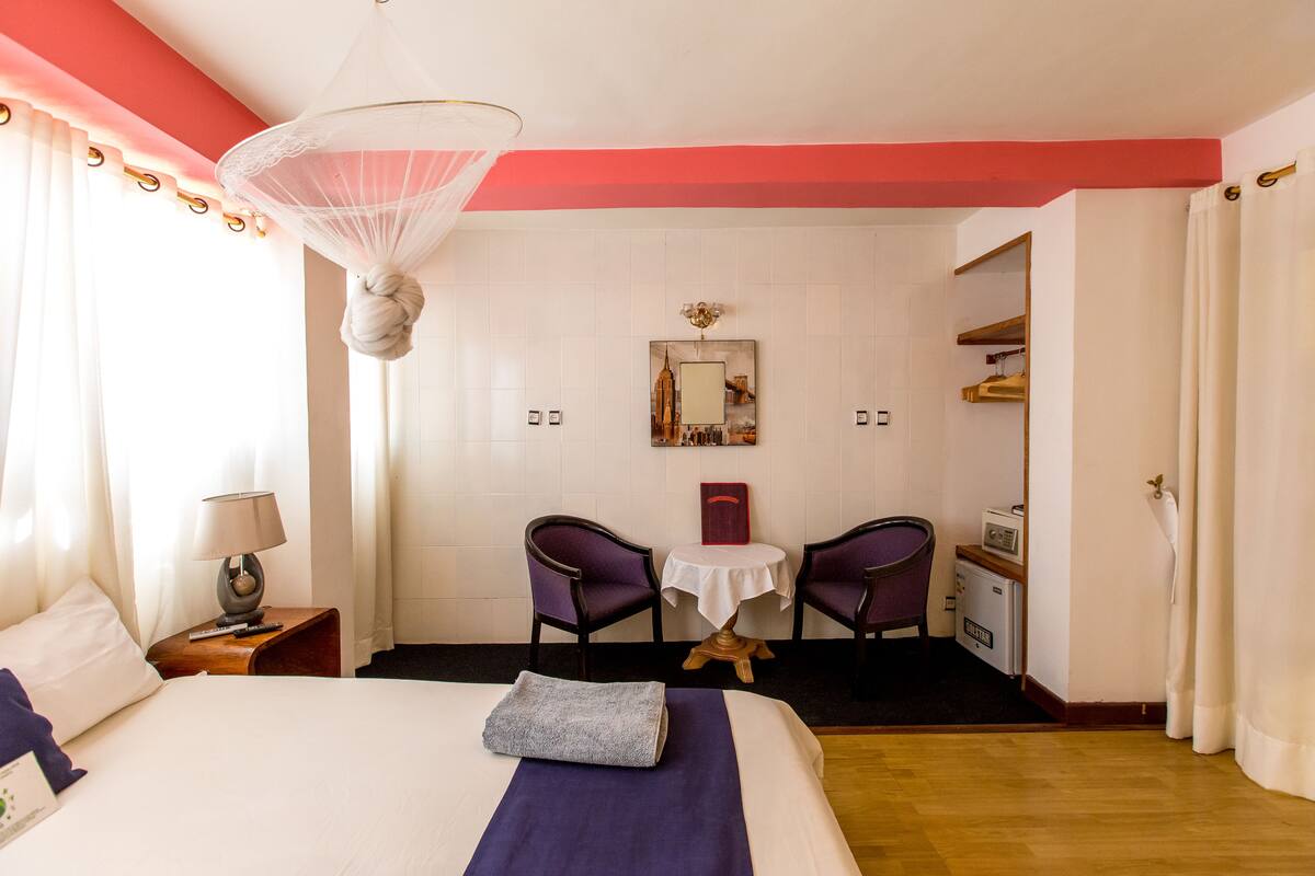 A hotel room with a bed, a bedside table with a lamp, a small table with chairs, a painting on the wall, and a wardrobe.