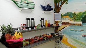 A breakfast bar with a tropical mural, a ship decoration, and a variety of fruits and beverages. - Pousada do Marinho (Búzios)
