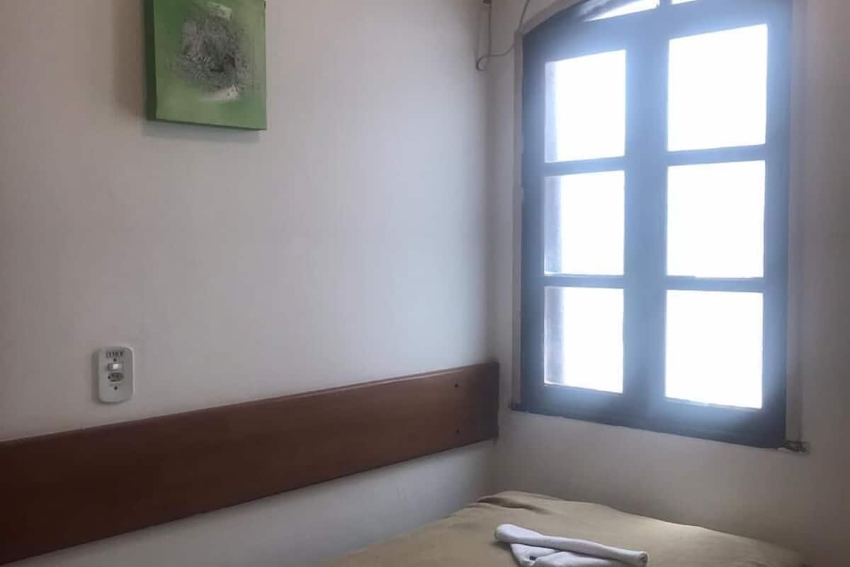 A small, single-bed room with a window, a green artwork on the wall, and a white air conditioning unit.