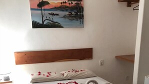 A hotel room with a bed, a painting of a sunset landscape, and rose petals scattered on the floor. - Pousada do Marinho (Búzios)