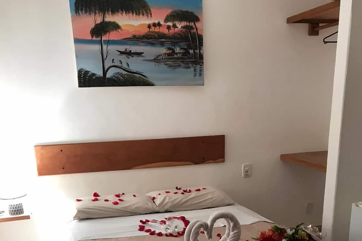 A hotel room with a bed, a painting of a sunset landscape, and rose petals scattered on the floor.