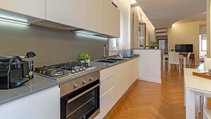 Een moderne keuken met een gasfornuis, oven en aanrechtbladen van roestvrij staal. Er is een eethoek met een tafel en stoelen zichtbaar op de achtergrond.