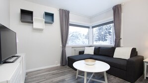 Een moderne woonkamer met een grijze bank, een witte salontafel en een flatscreen televisie op een wit kastje. Twee wandplanken, één grijs en één blauw.