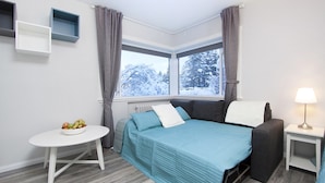 Een kleine kamer met een bed, een tafeltje met fruit, een nachtkastje en een raam met uitzicht op met sneeuw bedekte bomen.