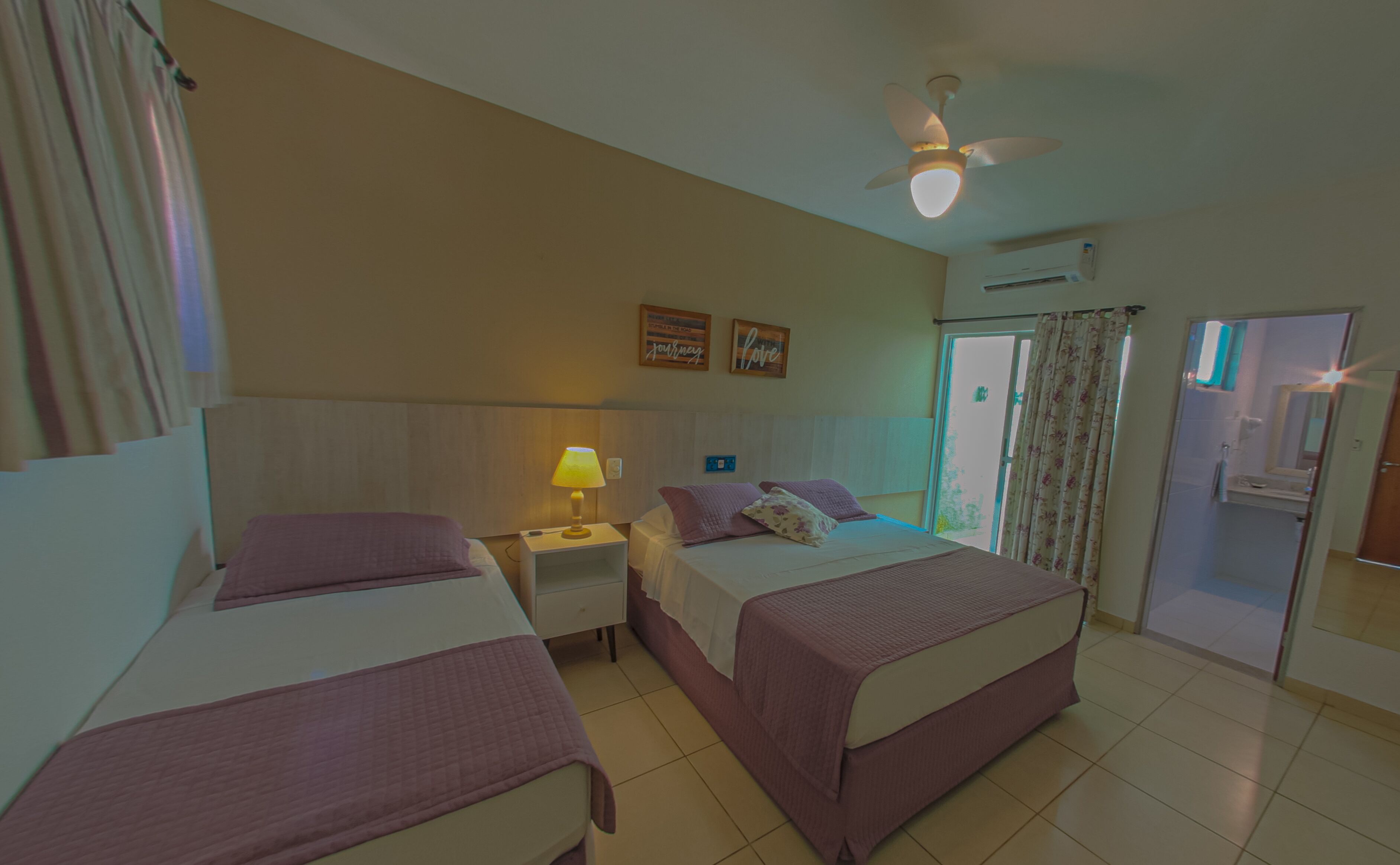 A hotel room with two beds, a ceiling fan, a nightstand with a lamp, and a bathroom visible through an open door.