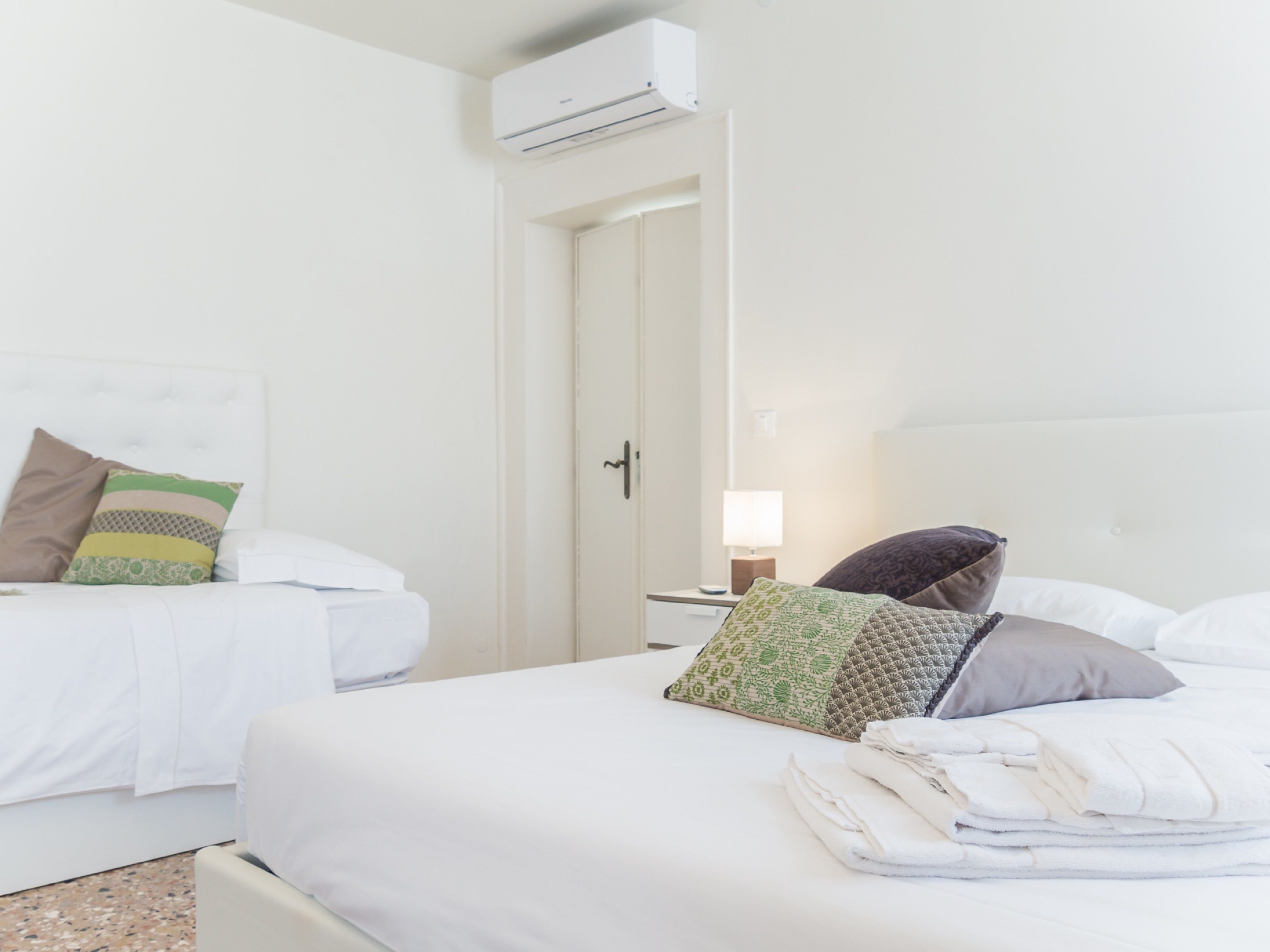 A hotel room with two beds, a door, an air conditioning unit, and a nightstand with a lamp.