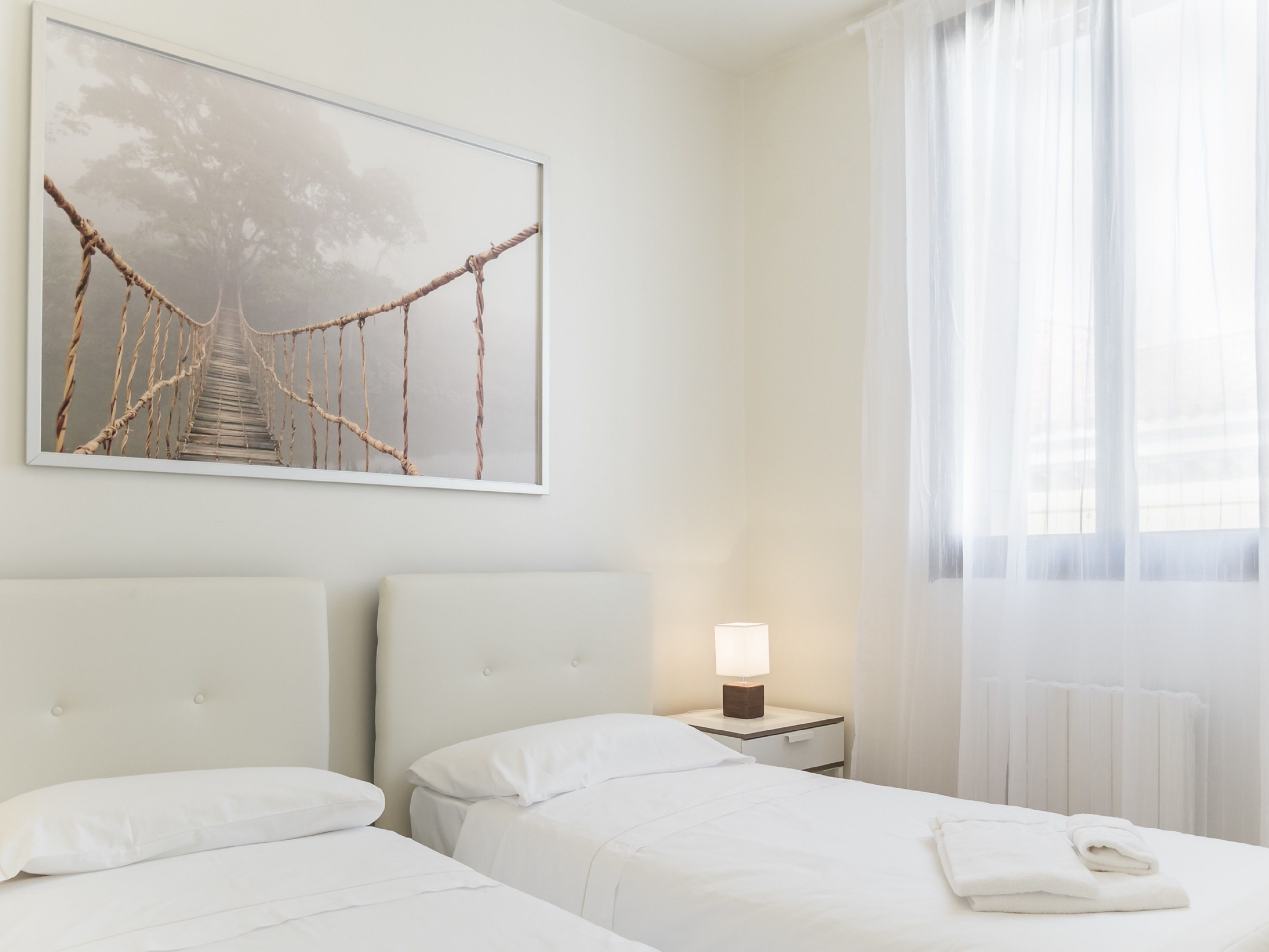Ein Hotelzimmer mit zwei Betten, einem Nachttisch mit Lampe, einem gerahmten Bild einer Brücke und einem großen Fenster mit durchsichtigen Vorhängen.