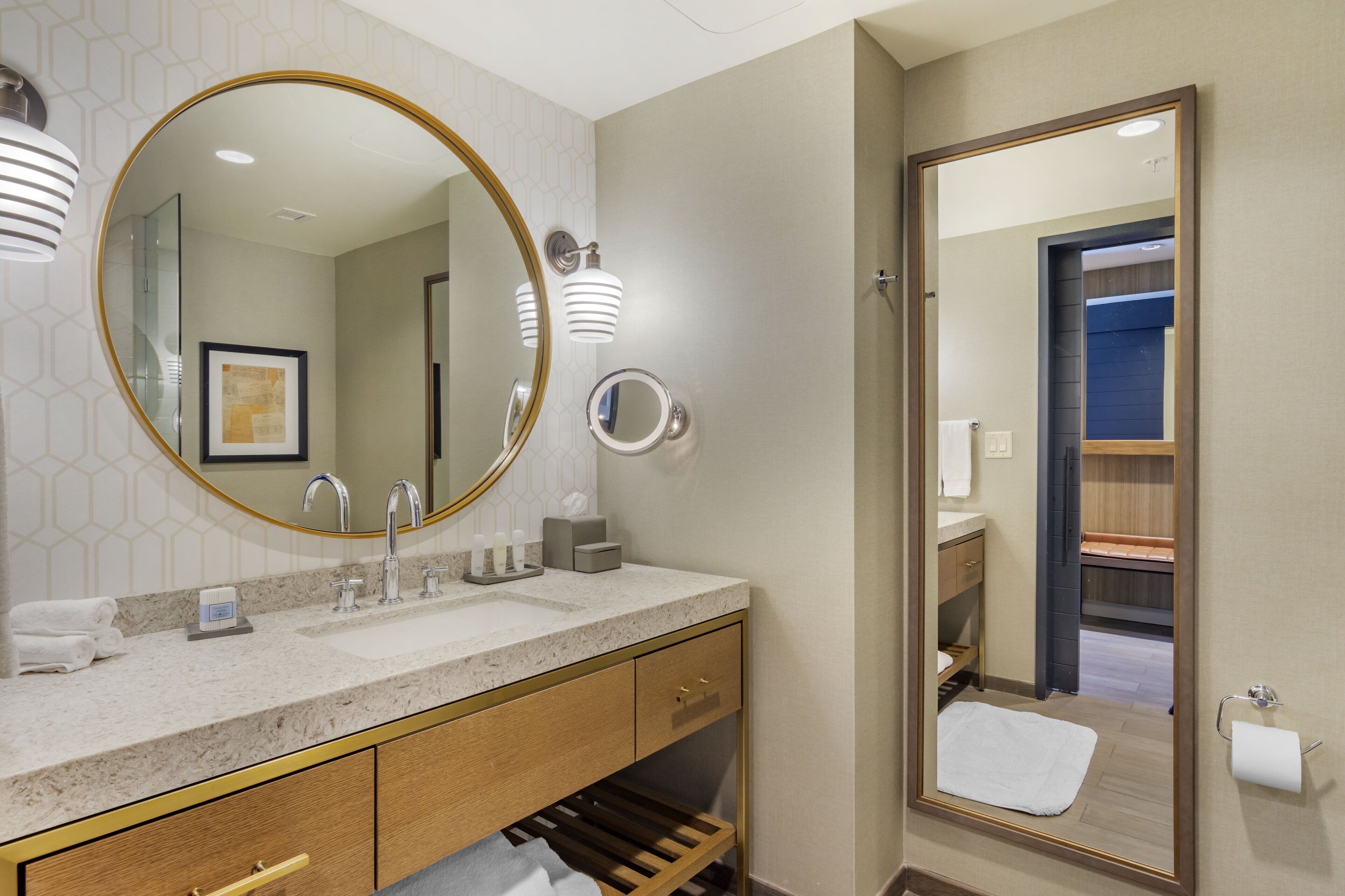 A modern bathroom with a large round mirror, a sink with a marble countertop, and a wooden vanity with drawers.
