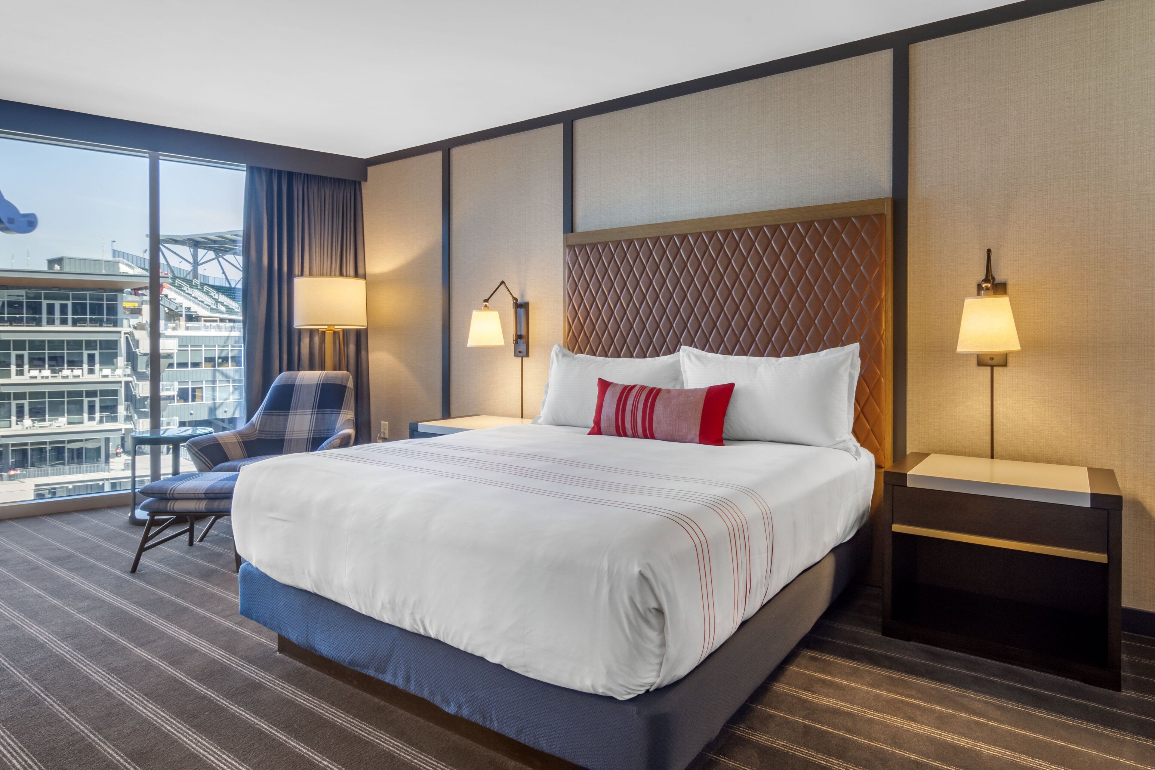 A hotel room with a large bed, two bedside tables with lamps, a chair, and a view of a cityscape.