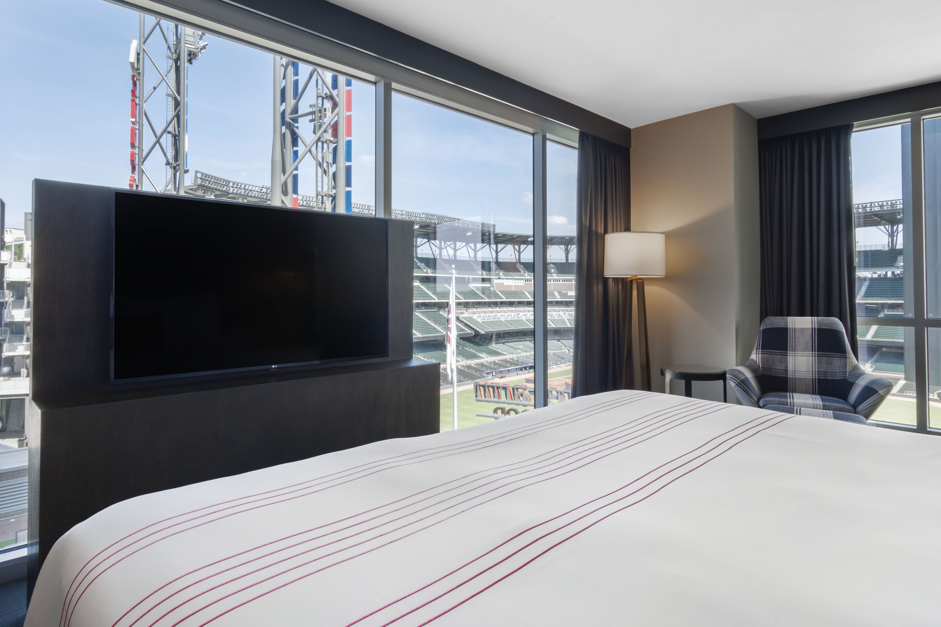 A hotel room with a large bed, a flat-screen TV mounted on the wall, a chair, and a view of a stadium through large windows.