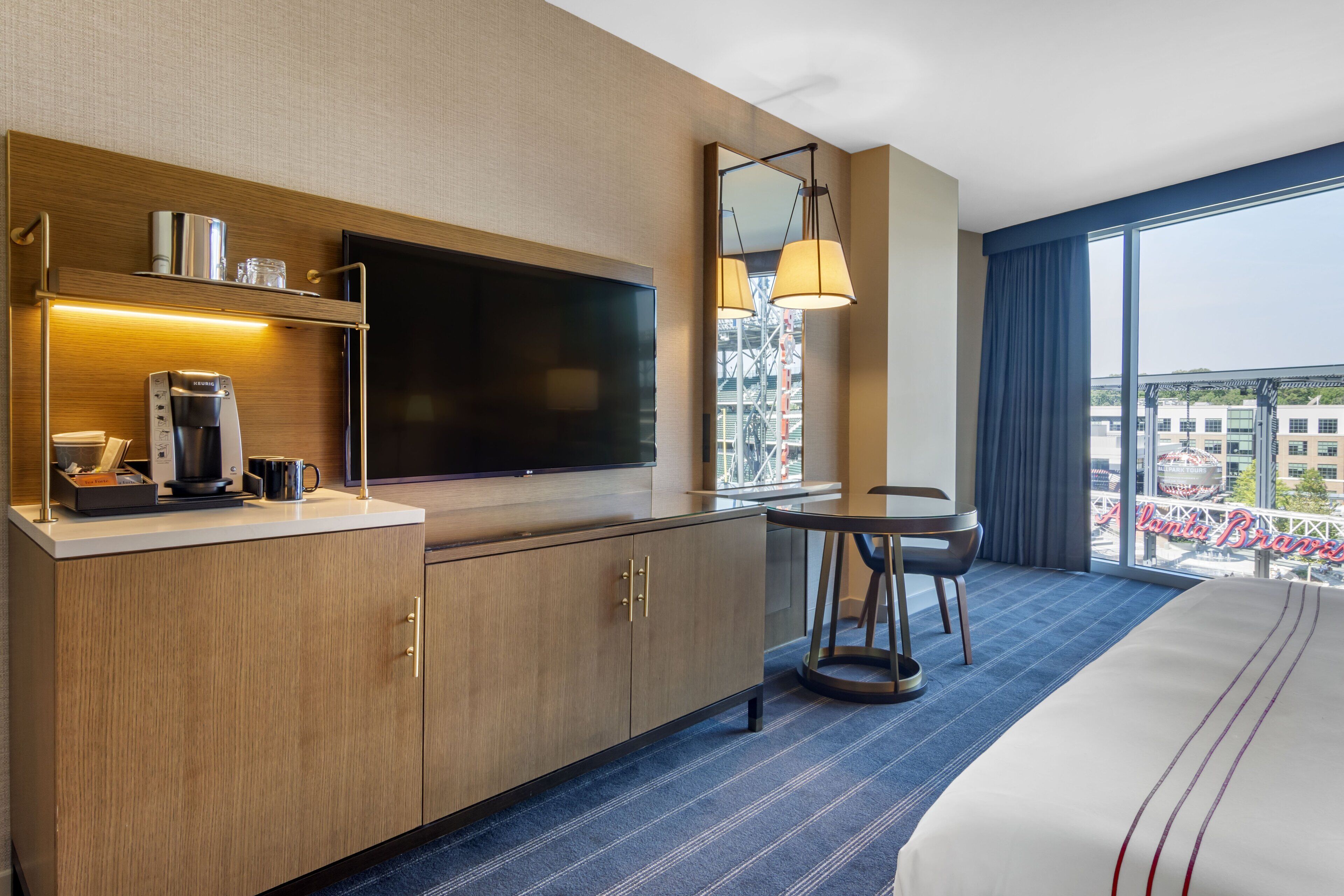 A hotel room with a large bed, a flat-screen TV, a coffee machine, a small table with a chair, and a large window with a view of the city.