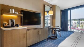 A hotel room with a large bed, a flat-screen TV, a coffee machine, a small table with a chair, and a large window with a view of the city.