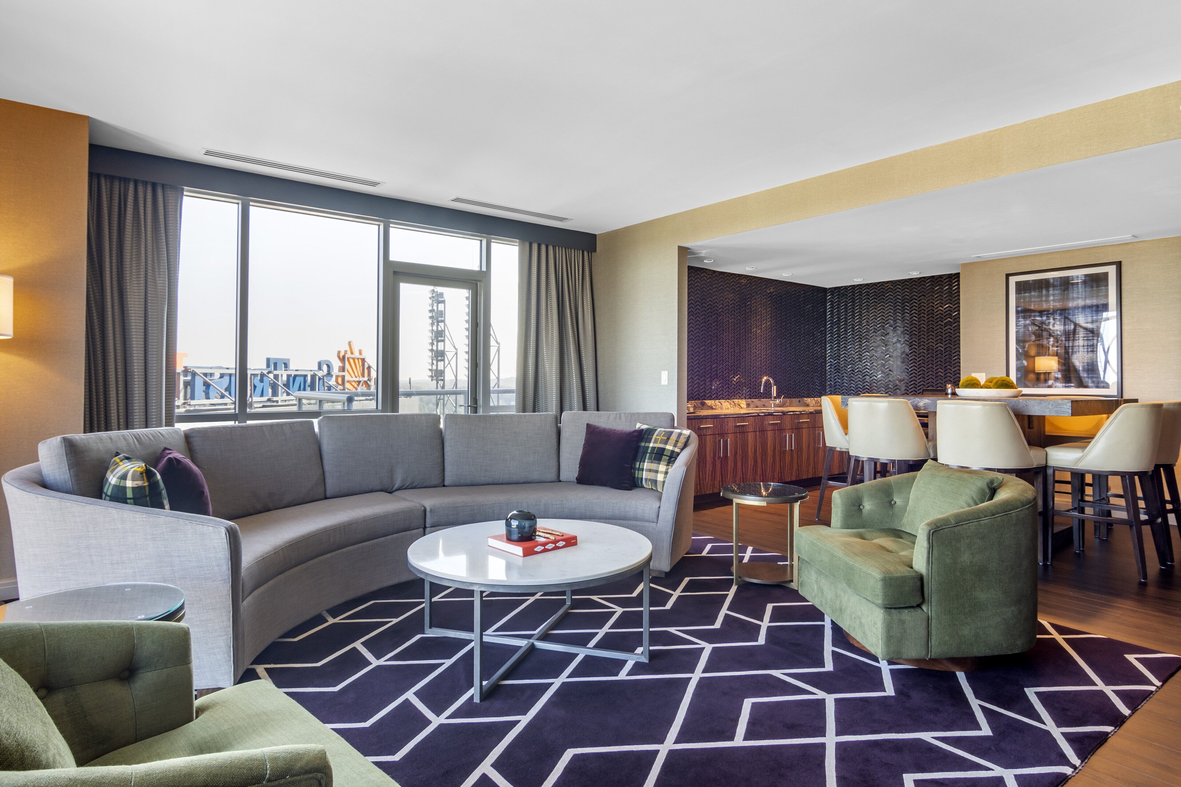 A modern hotel room with a curved gray sofa, a green armchair, a round coffee table, and a dining area with a kitchen in the background.