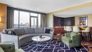 A modern hotel room with a curved gray sofa, a green armchair, a round coffee table, and a dining area with a kitchen in the background.