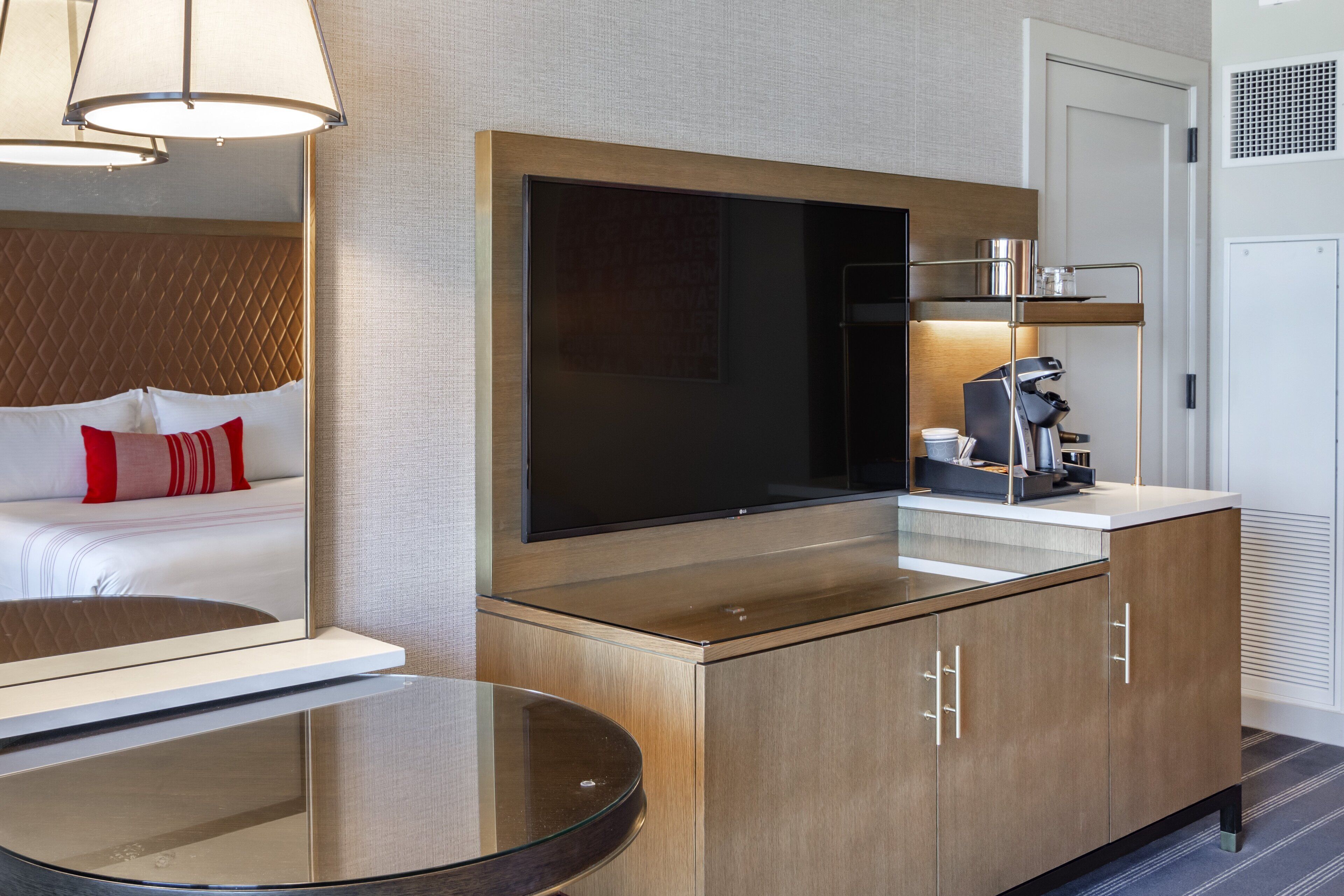 A modern hotel room with a large flat-screen TV mounted on a wooden cabinet, a bedside table with a glass top, and a coffee machine on a white countertop.