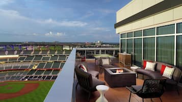 A rooftop terrace with a view of a baseball stadium, furnished with sofas, chairs, and a fire pit.