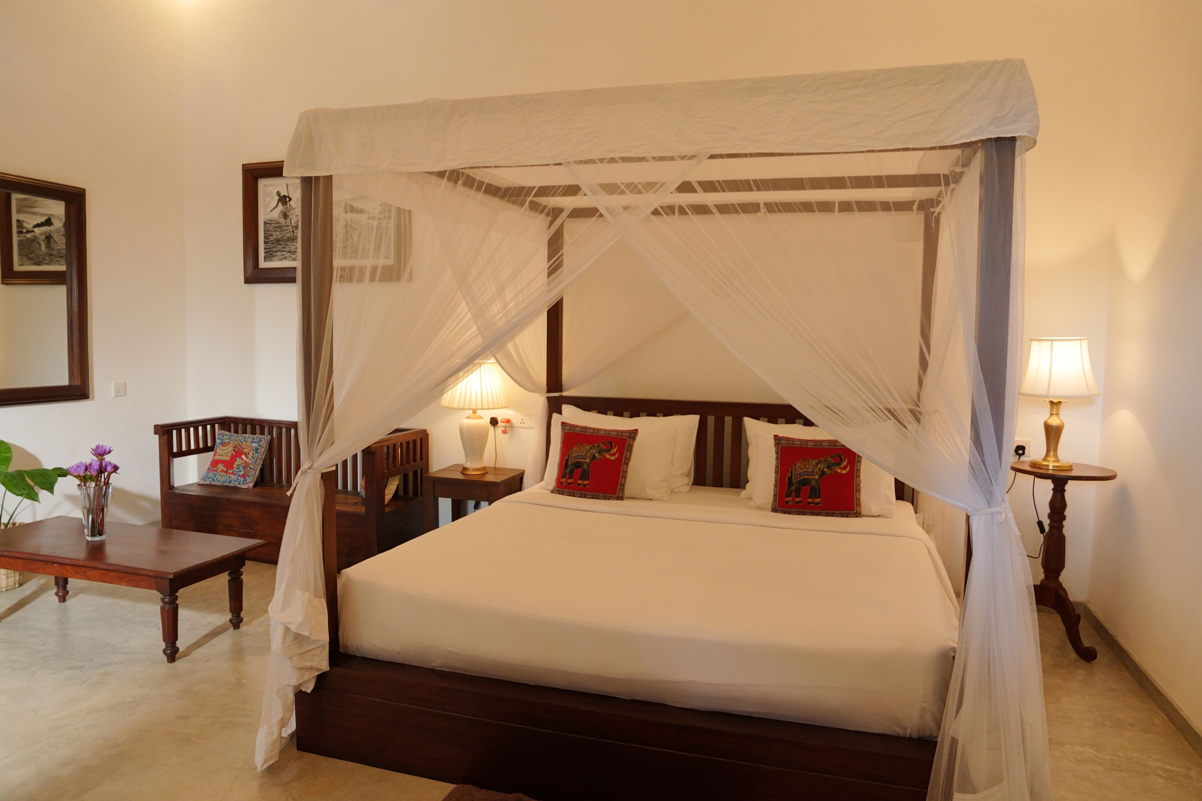 A four-poster canopy bed with white bedding and decorative pillows, a bedside table with a lamp, and a small table with a vase of flowers.