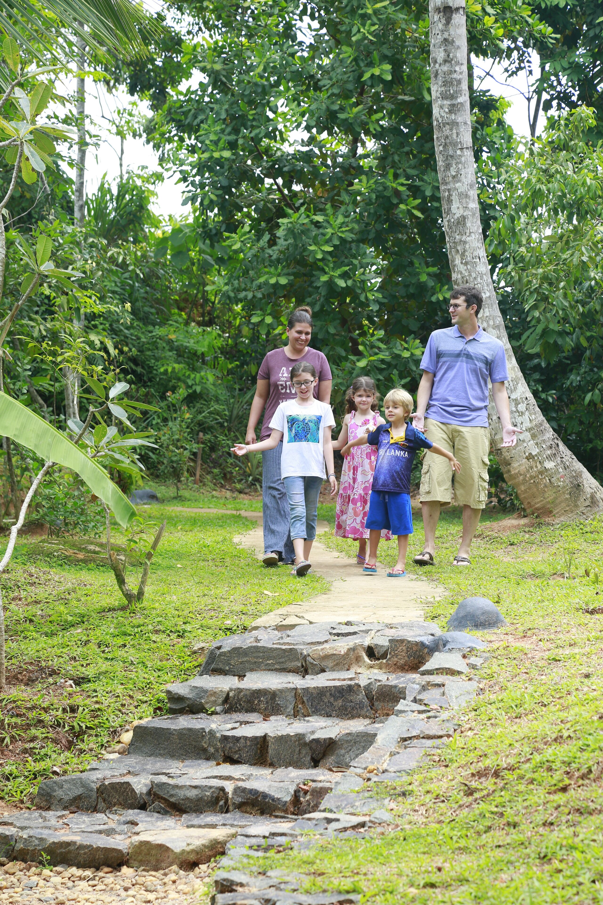 Une famille se promène sur un sentier de pierres dans un espace verdoyant et luxuriant, avec des arbres et un grand tronc d’arbre sur la droite.