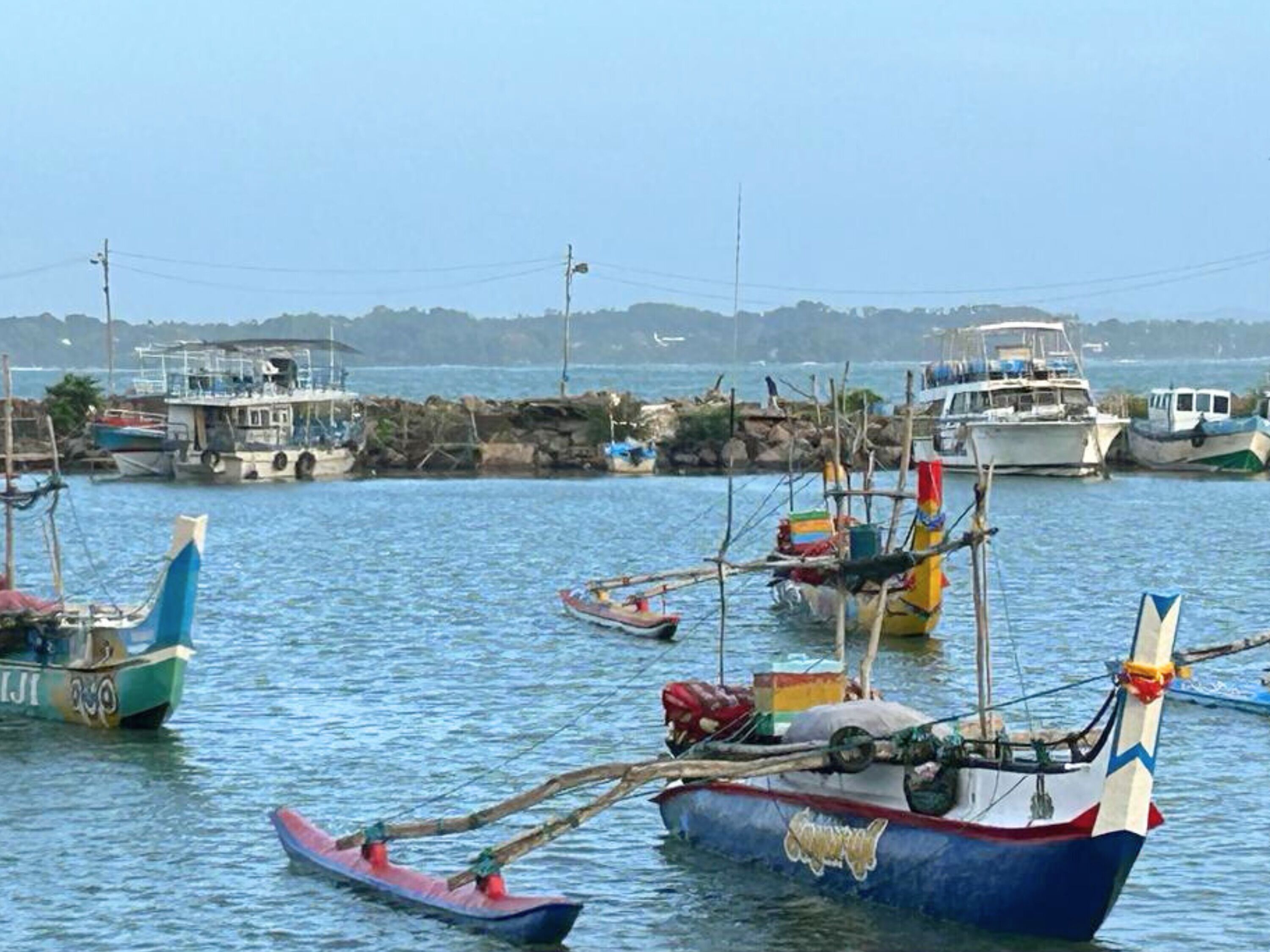 A harbor with various boats docked, including a blue and white boat with a flag, a smaller red boat, and a white boat with a cabin.