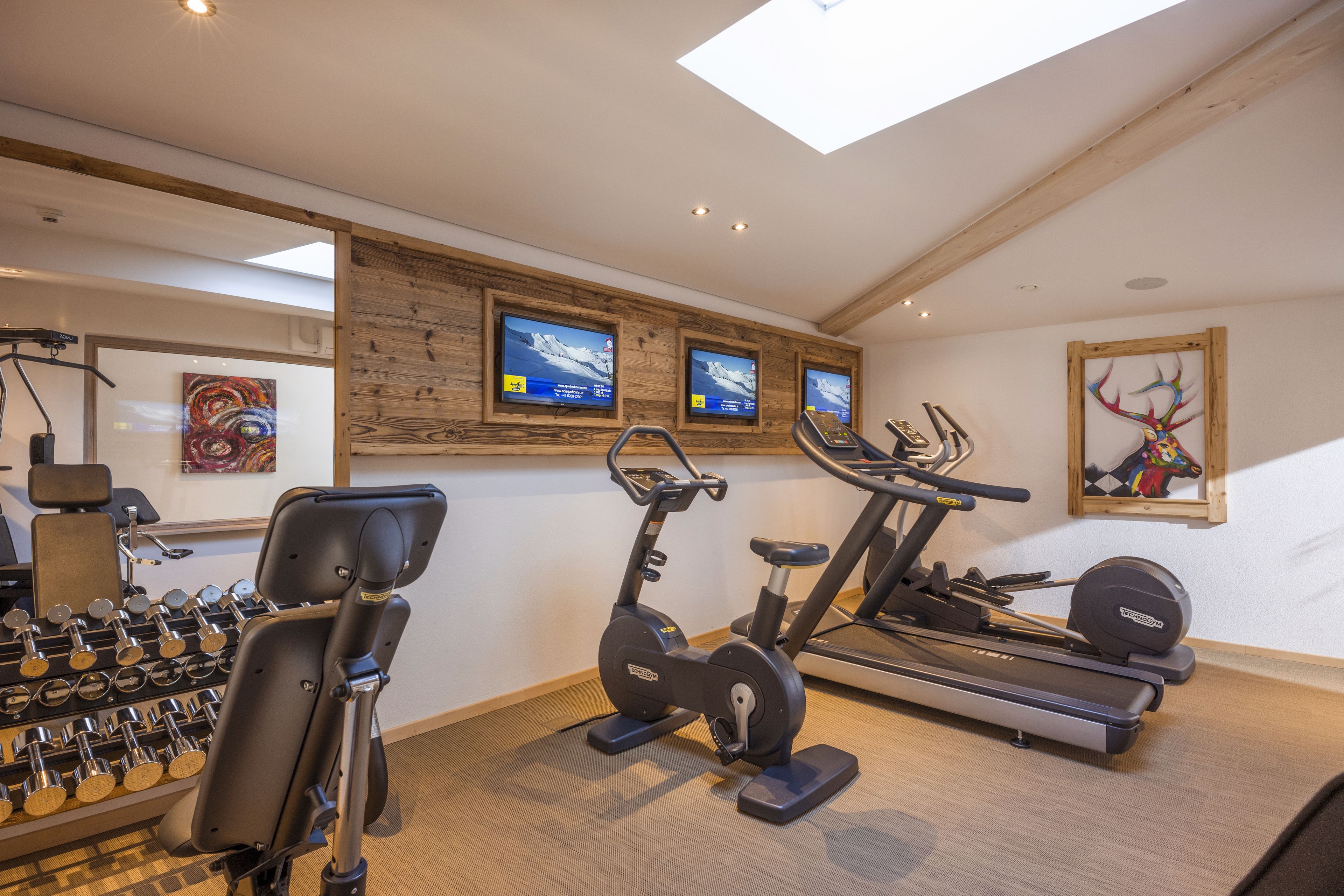 A gym with exercise equipment, including a rowing machine, an elliptical, and a treadmill. There are three TV screens on the wall displaying content.