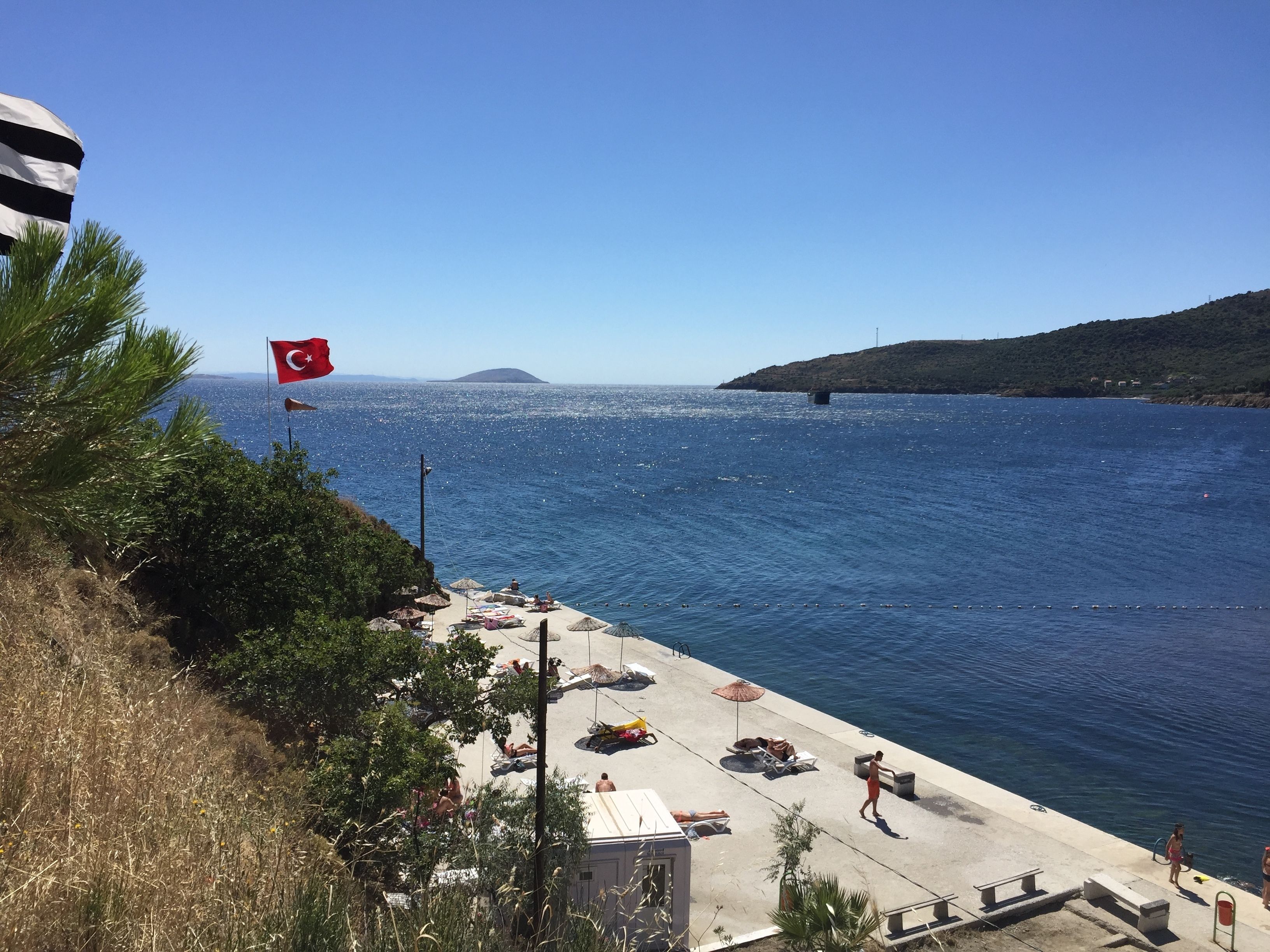 A coastal area with a flag, beach umbrellas, lounge chairs, and a clear view of the sea.